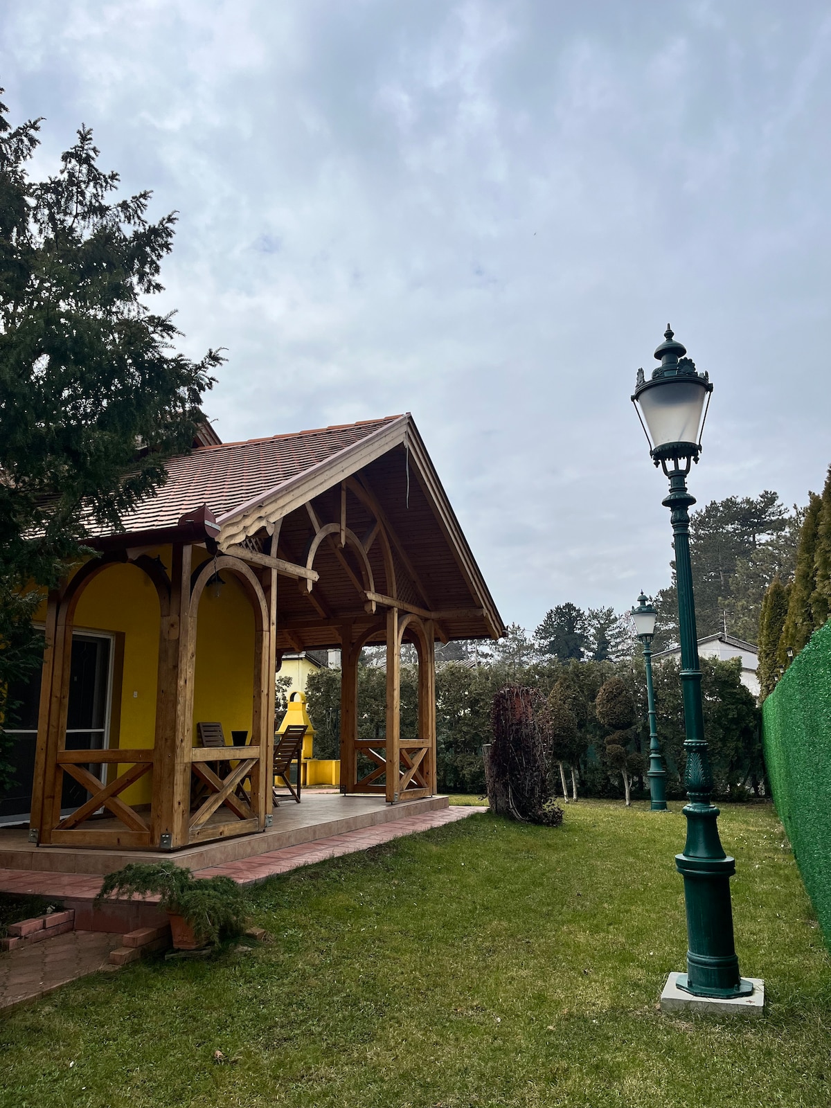 A wooden gazebo with an arched roof is set in a grassy area, offering a space to relax outdoors. Nearby, a decorative lantern stands on a pathway, adding a classic touch to the surroundings. Lush greenery provides a natural backdrop.