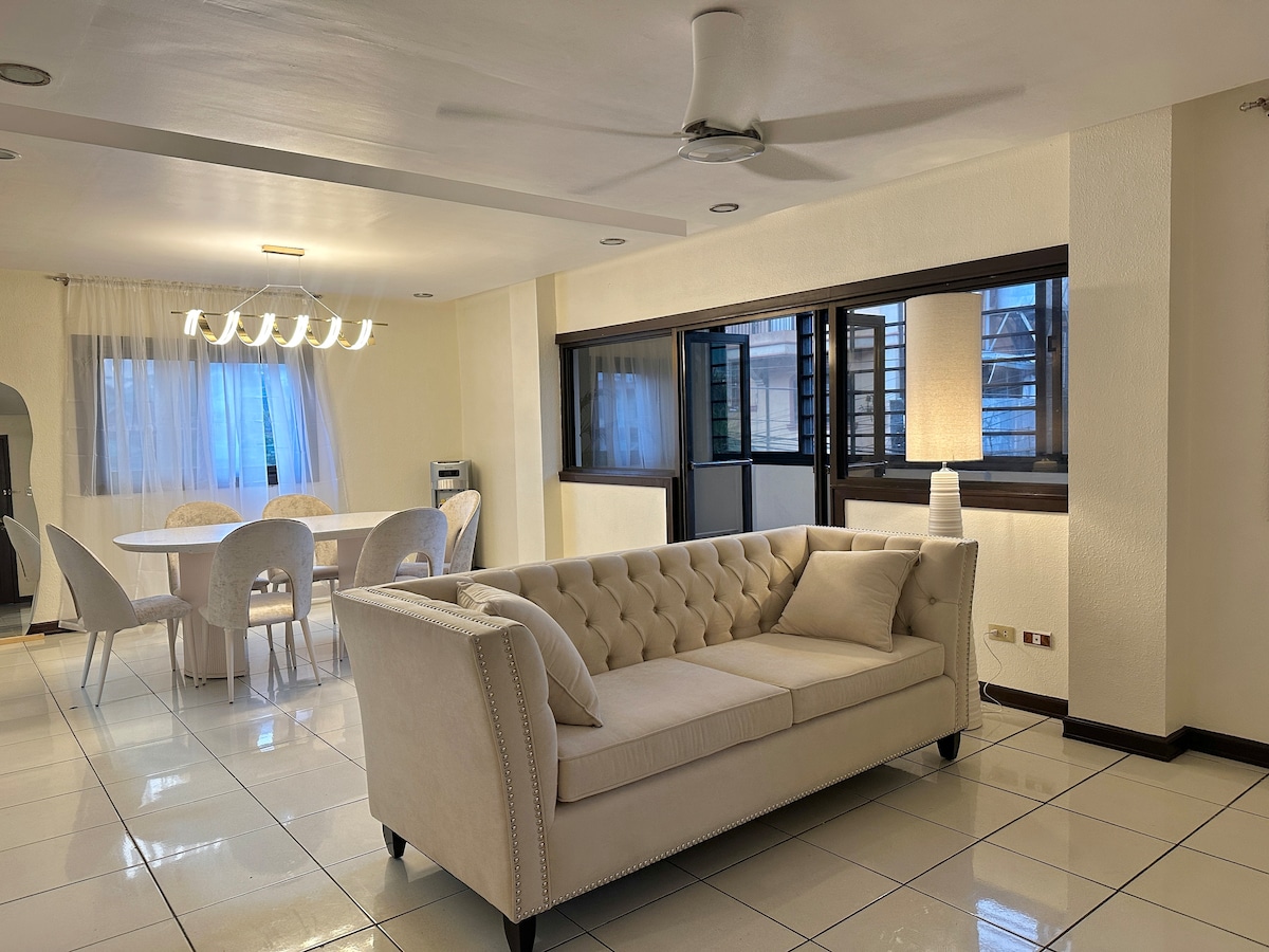 A spacious living area features a light-colored tufted sofa positioned next to large windows, allowing natural light to fill the room. A dining table with six upholstered chairs can be seen in the background, enhancing the open layout of the space.