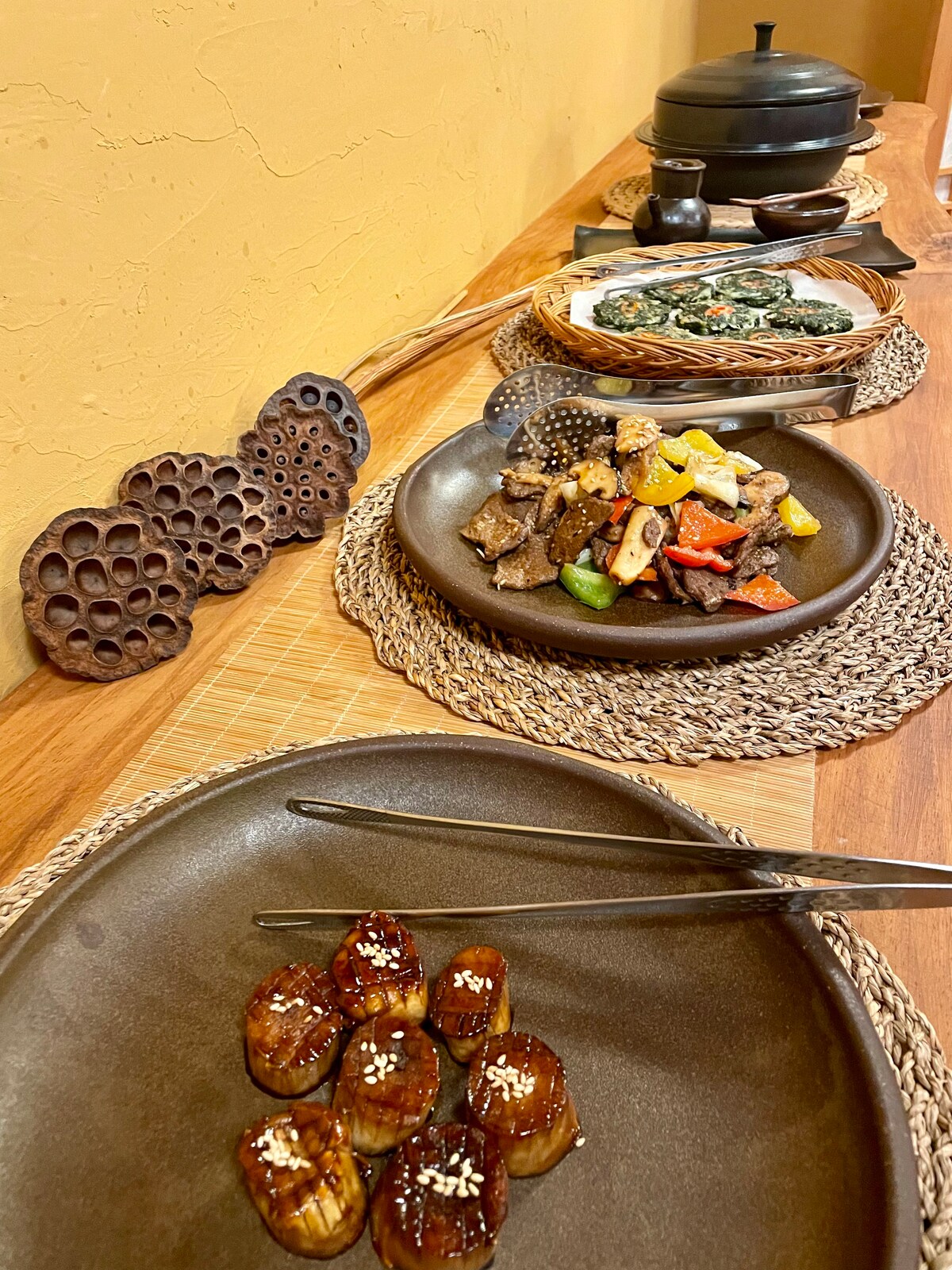 A wooden dining table is set with various dishes showcasing colorful vegetables and unique culinary presentations. A plate of glistening roasted eggplant garnished with sesame sits prominently, accompanied by a selection of stir-fried vegetables. Natural woven placemats add texture to the arrangement.