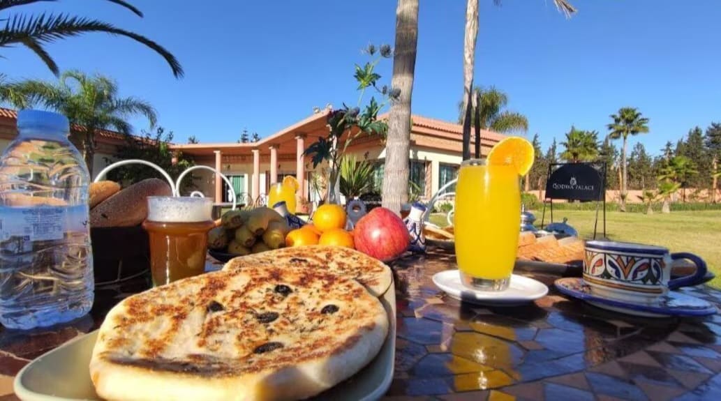 A table is set outdoors, displaying a variety of breakfast items including flatbreads, fresh fruit, and a glass of orange juice. A scenic view of palm trees and the property can be seen in the background.