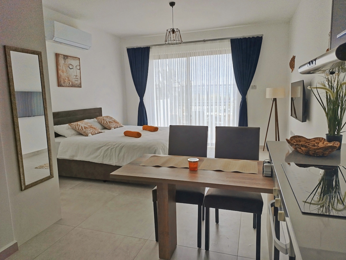 A spacious studio apartment features a comfortable bed with orange accent pillows and large windows covered by blue drapes. A wooden dining table with two chairs is positioned nearby, and a wall-mounted television is visible. Soft natural light fills the room, highlighting the neutral decor.