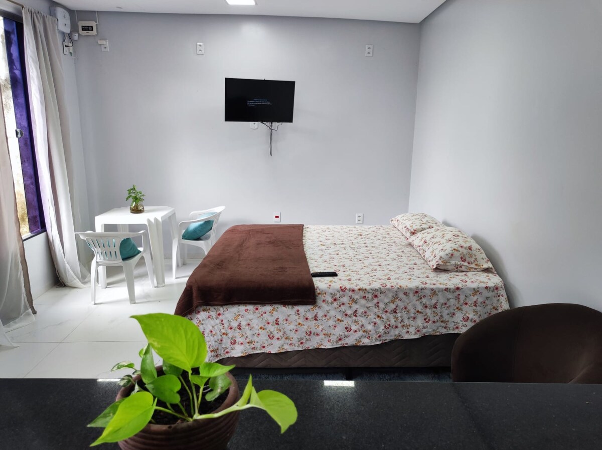 A comfortable sleeping area is visible, featuring a decorative bed with floral bedding. A small white table and two chairs are positioned nearby, accompanied by a green plant. A television is mounted on the wall, and natural light enters through a window covered by sheer curtains.