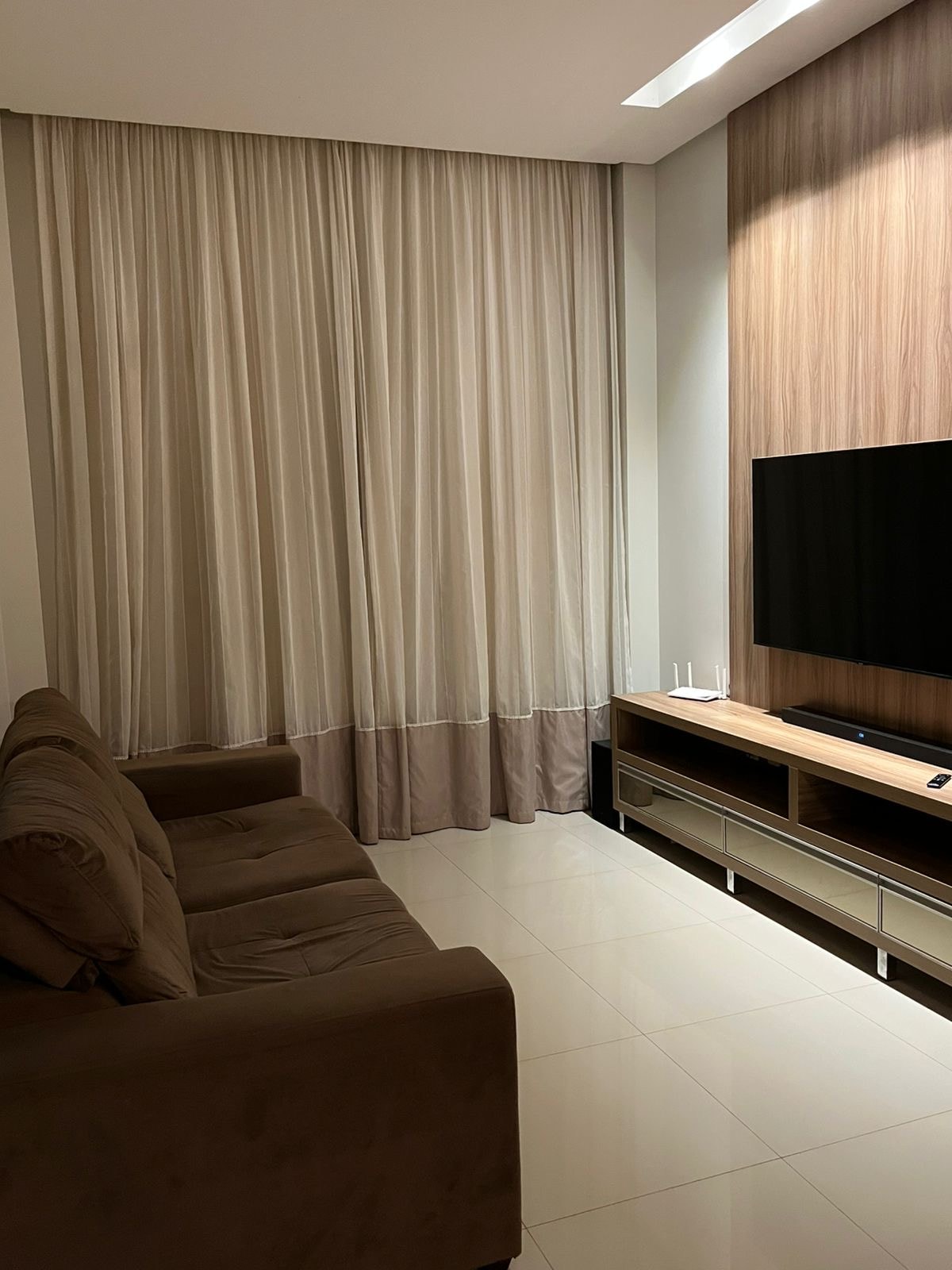 A living area is presented with a brown sofa facing a wall-mounted television. Light-colored curtains drape over a large window, allowing natural light to filter in. A low wooden media console sits below the TV, creating a balanced and functional space.