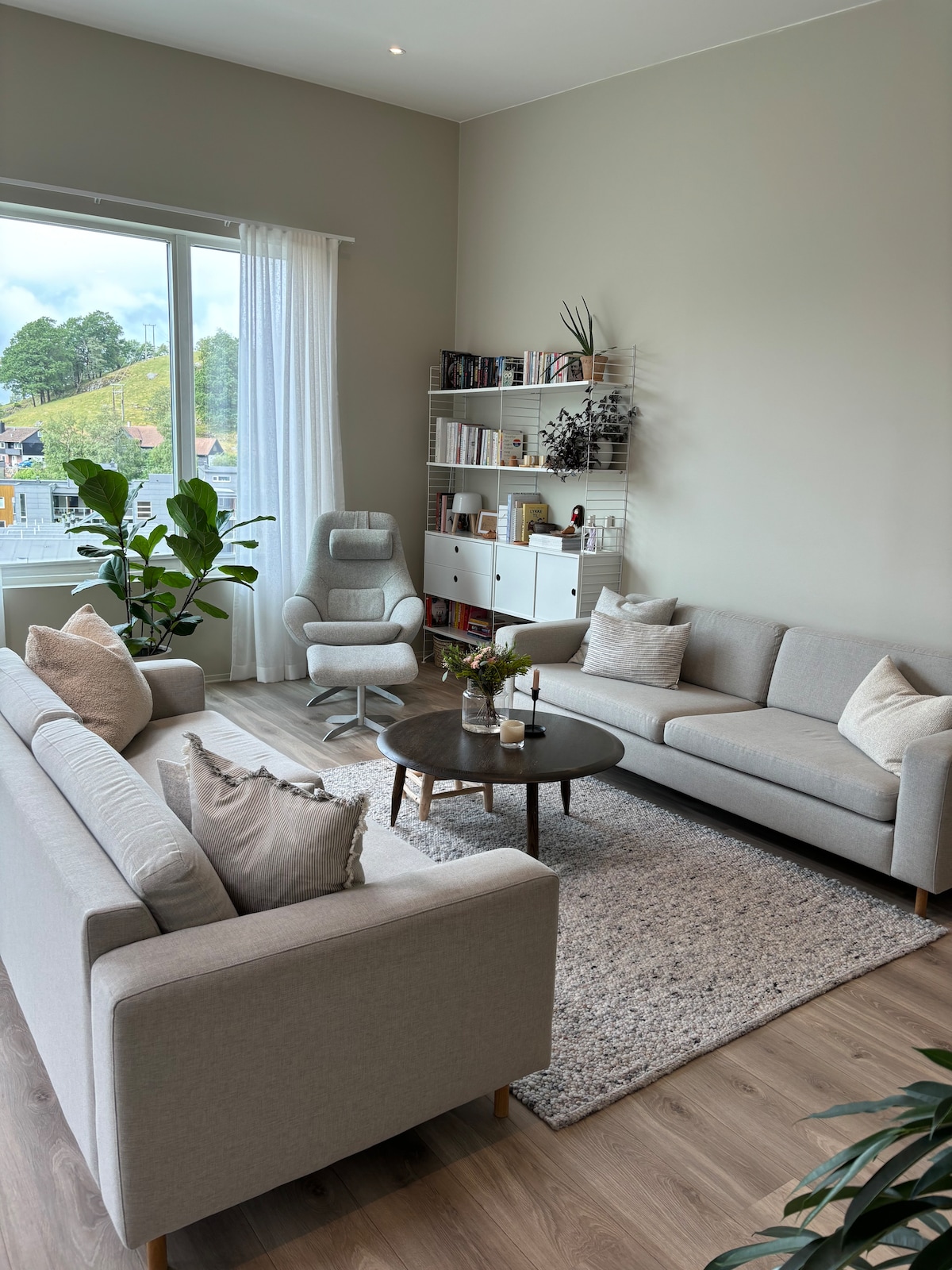 The living area is furnished with two light-colored sofas and a round coffee table at the center. A modern armchair sits beside a large window, allowing natural light to brighten the space. Shelving units occupy the wall, displaying books and decorative items, complemented by indoor plants.