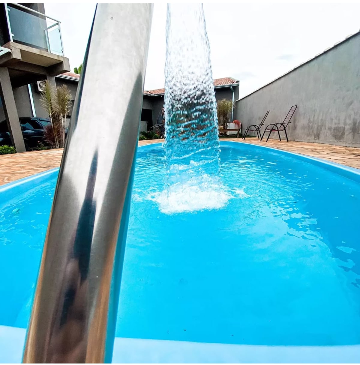 A sparkling blue pool is shown with water cascading from a stainless steel feature. The surrounding area is paved, and nearby, two chairs are positioned against a gray wall, with greenery visible in the background.