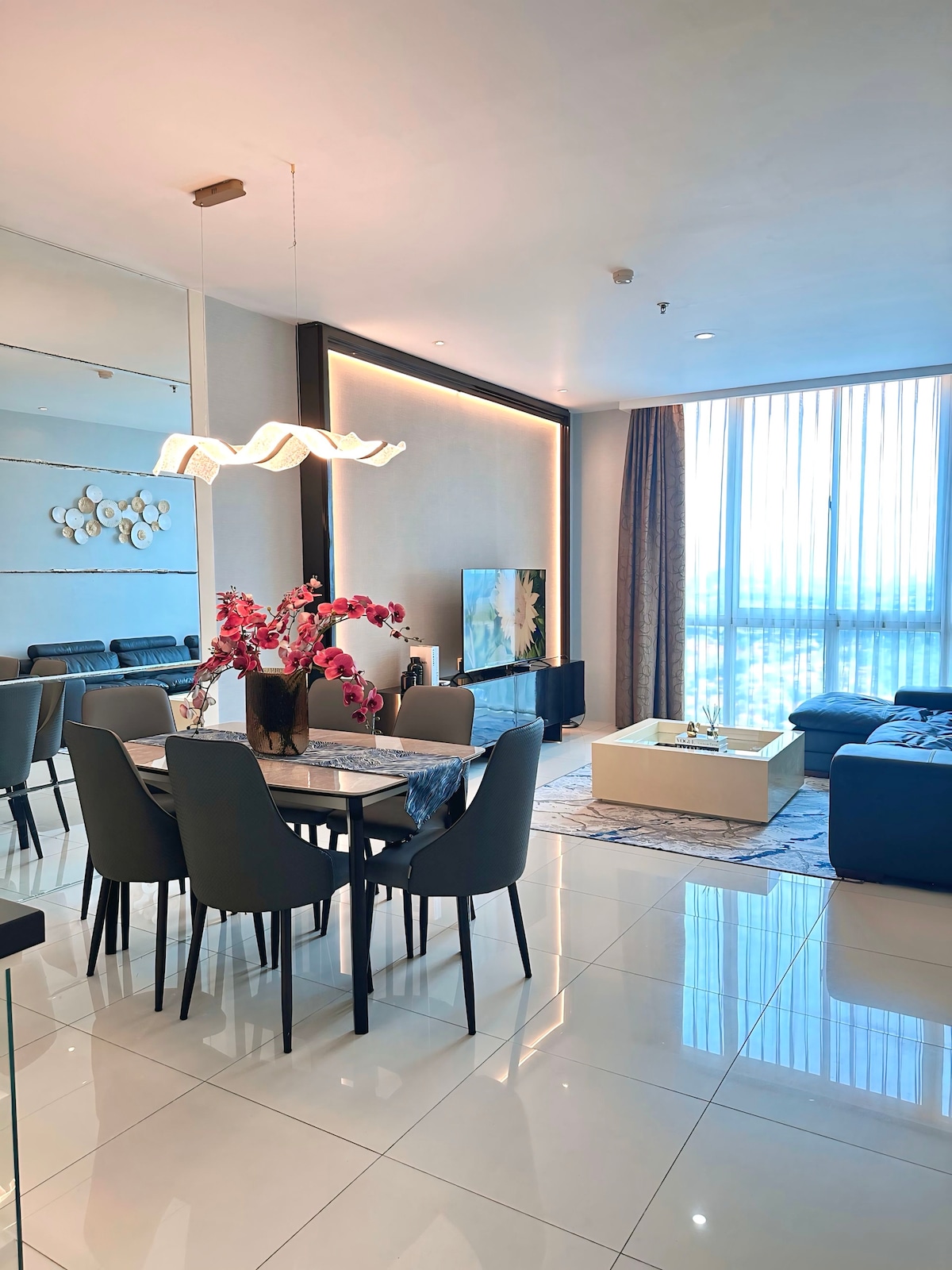 A modern dining area is shown with a dark wooden table surrounded by grey upholstered chairs. An artistic light fixture hangs above. A comfortable seating arrangement with a dark sofa is visible in the background, complemented by floor-to-ceiling windows that allow natural light to fill the space.