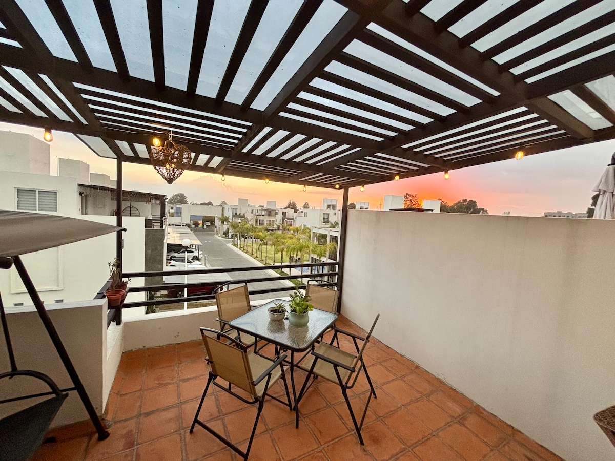 A terrace is presented with a structured pergola, featuring a table set for four and surrounded by comfortable chairs. Ambient lighting is seen overhead, and potted plants add a touch of greenery. The horizon displays a subtle sunset, enhancing the serene atmosphere.