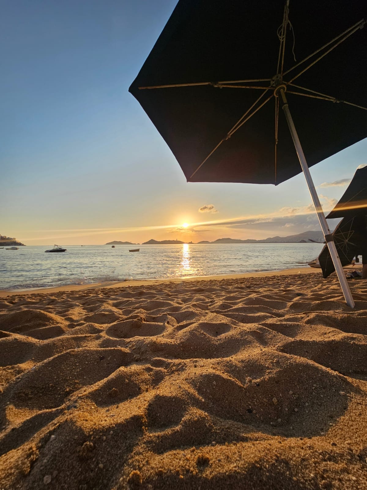 The image features a sandy beach with gentle waves lapping at the shore. An umbrella is positioned in the foreground, providing shade. The sun is setting on the horizon, casting a warm glow over the water and creating a serene atmosphere.