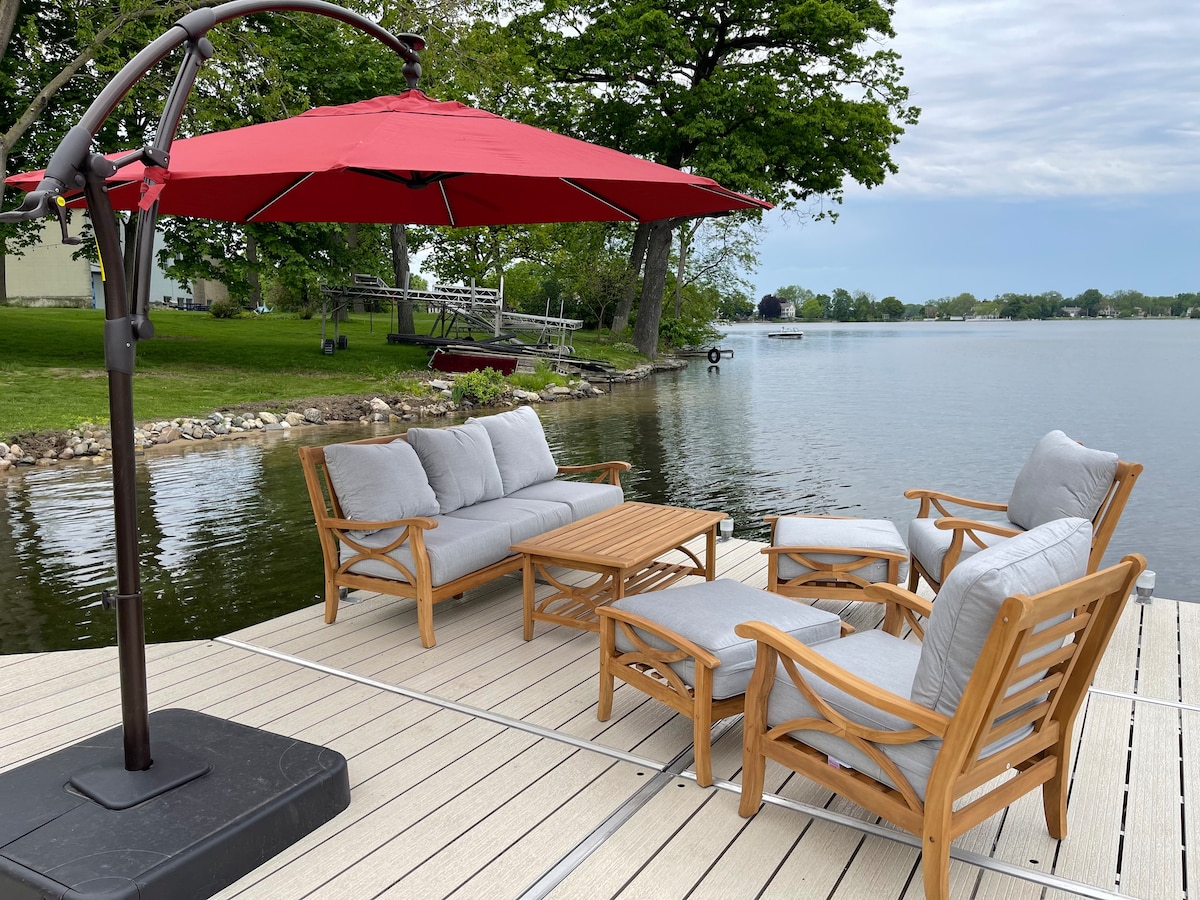 A water-side seating area features light-colored cushioned furniture arranged around a wooden coffee table. A red umbrella provides shade, complementing the serene lake view lined with greenery. The calm water reflects the surroundings, extending the outdoor relaxation space.