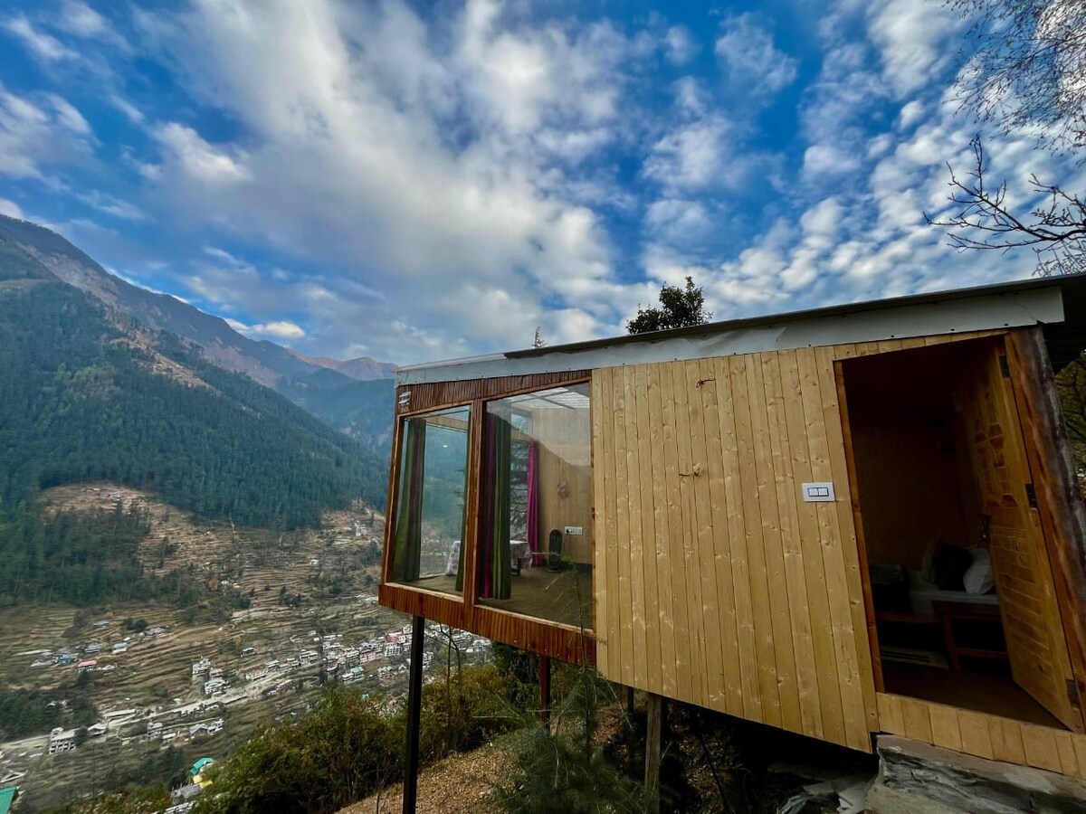 A contemporary tree house design is showcased with wooden exterior walls and large glass windows, offering expansive views of the surrounding mountains and valley. A clear sky with scattered clouds enhances the tranquil setting. The entrance features a simple wooden door, inviting guests indoors.