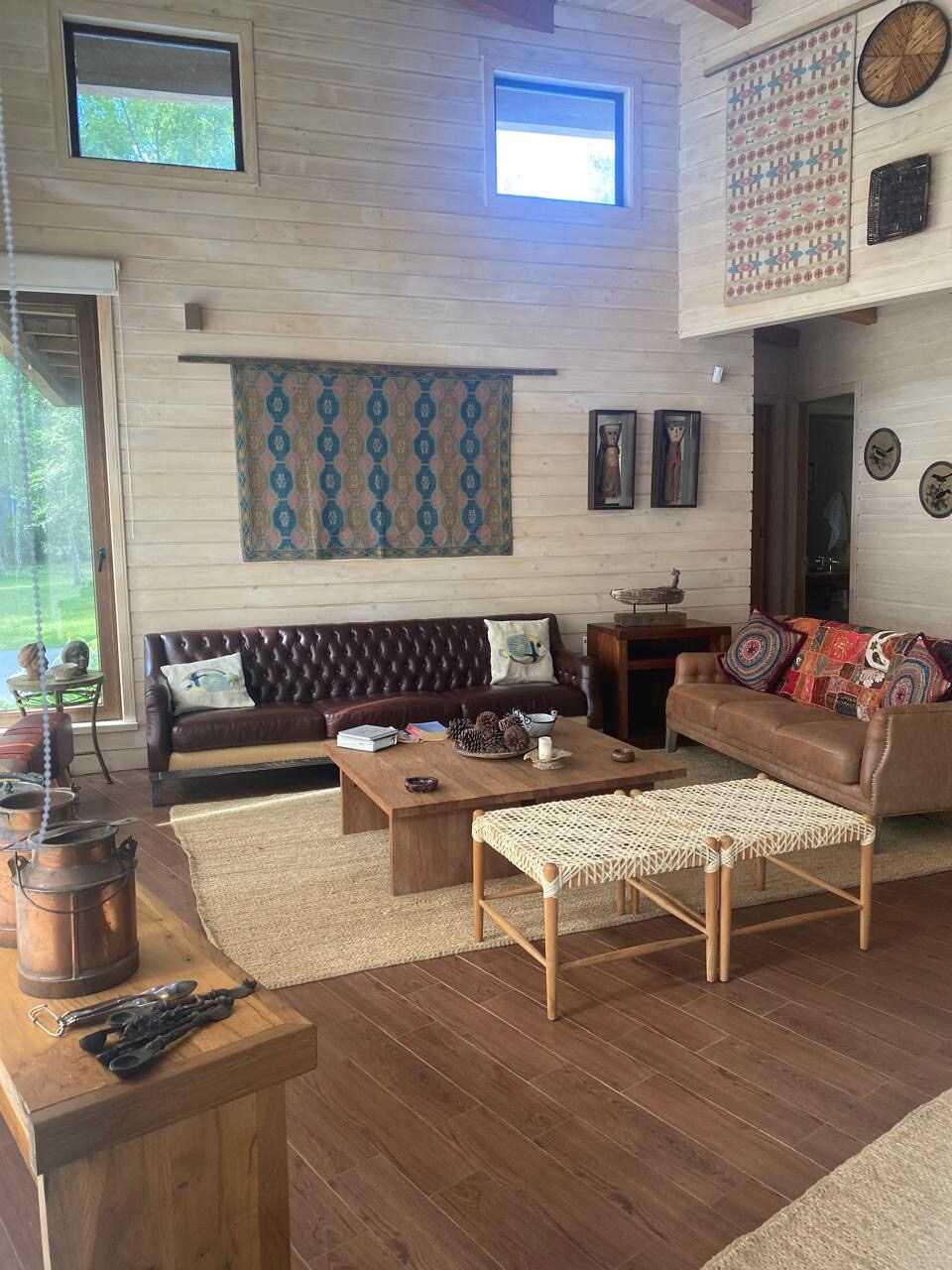 A spacious living area features a leather sofa and a cozy couch adorned with colorful cushions. A wooden coffee table occupies the center, surrounded by decorative wall hangings. Natural light enters through large windows, enhancing the warm wooden tones of the interior.