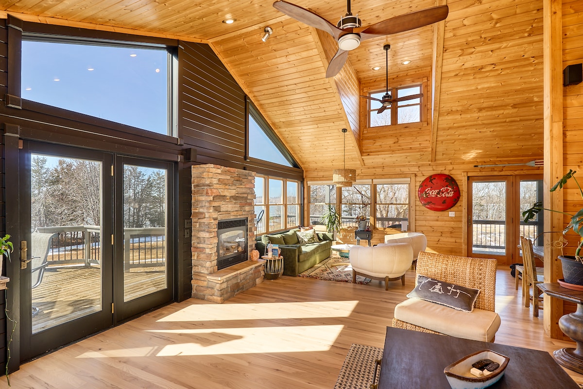 An open living area features a high vaulted ceiling and large windows that allow natural light to fill the space. A stone fireplace is positioned against a wooden wall, while comfortable seating is arranged around it. A ceiling fan hangs above, enhancing the airy atmosphere.