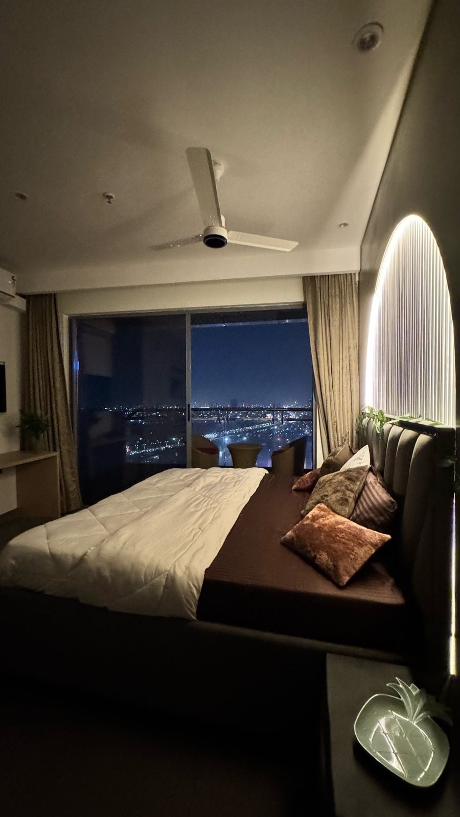 A cozy bedroom features a spacious bed adorned with decorative pillows. A large window offers expansive views of the city skyline at night, illuminated by city lights. Soft lighting accents the room, creating a calming atmosphere.
