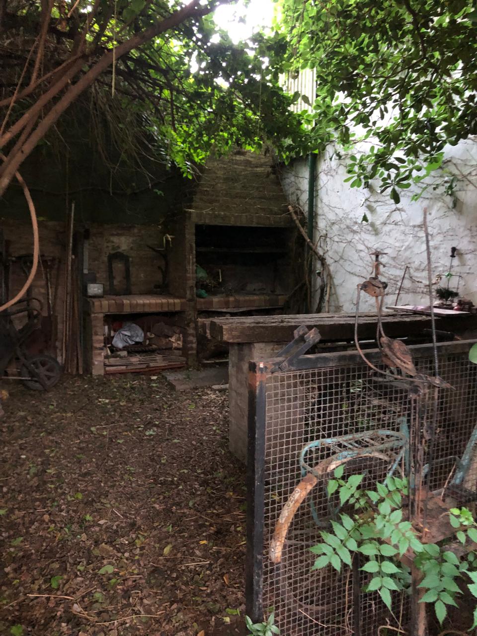 An outdoor space is showcased, featuring a stone structure with a fireplace against a rustic wall. Surrounding greenery creates a shaded area, while tools and equipment are organized on the ground. Natural elements blend with the curated features of the environment.