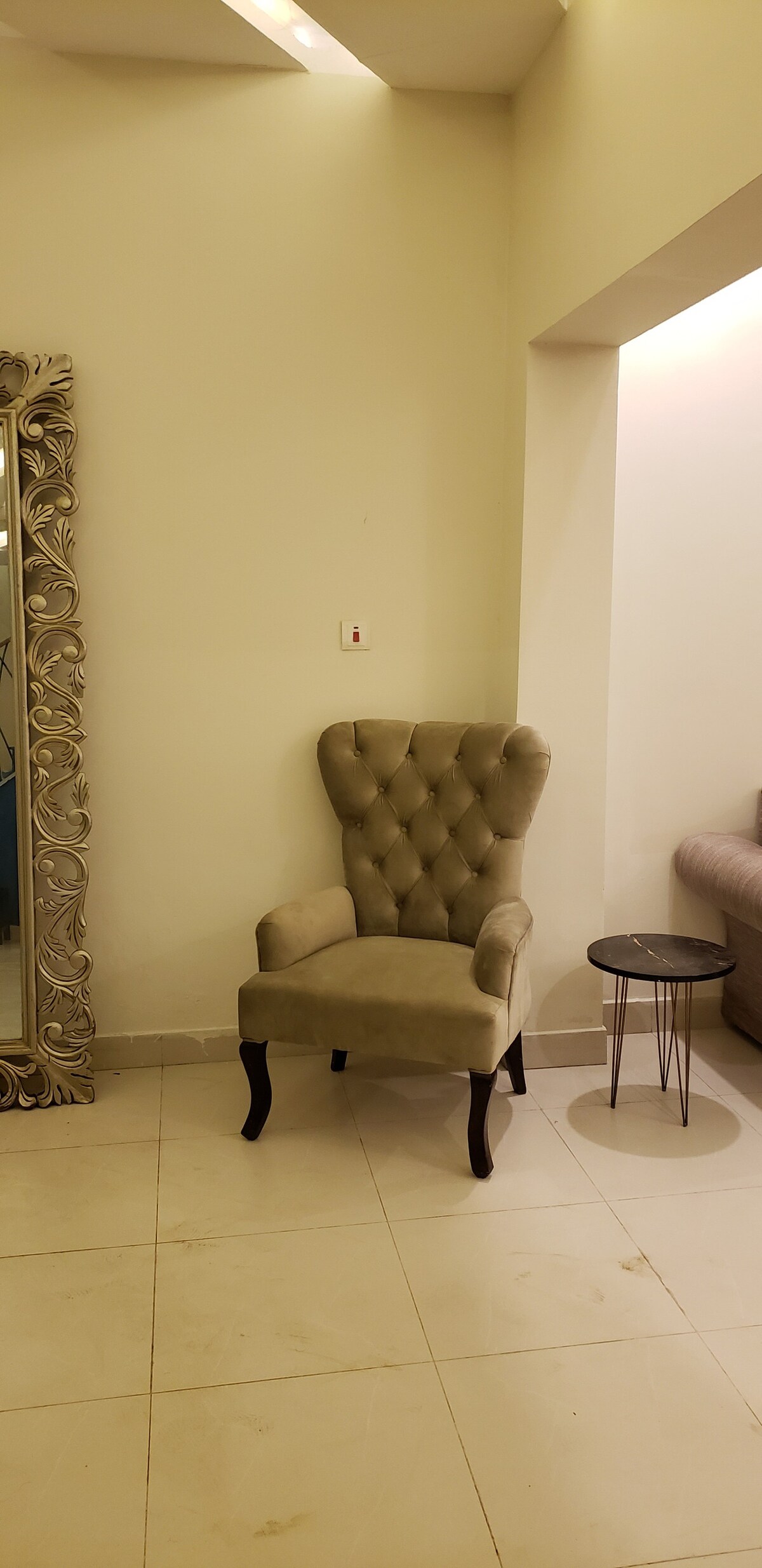 A tufted armchair with elegant detailing is positioned next to a small round table. The walls are painted in a light tone, enhancing the overall brightness of the space. A large, decorative mirror features intricate designs and reflects natural light.