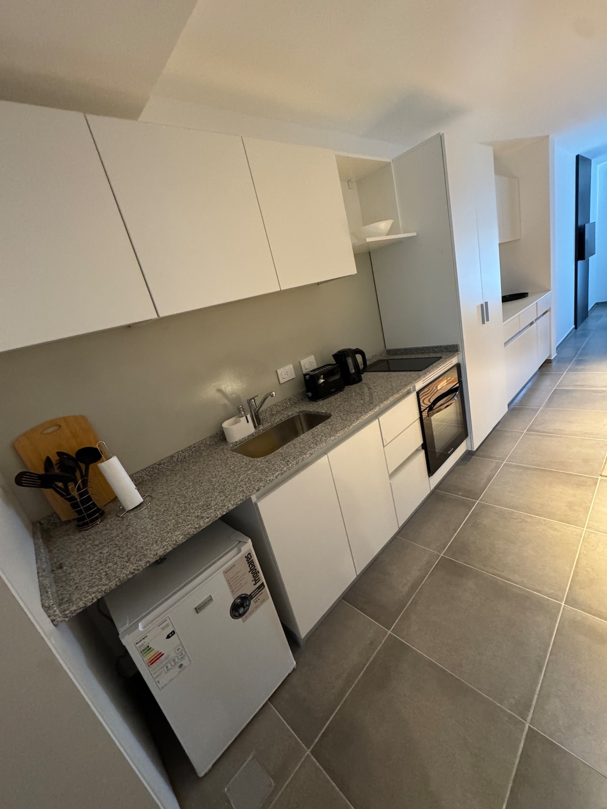 A modern kitchen area features sleek white cabinetry and a gray countertop. Essential appliances include a refrigerator, kettle, and oven. Natural light is provided by a nearby window, illuminating the tiled floor while creating a clean and functional space.