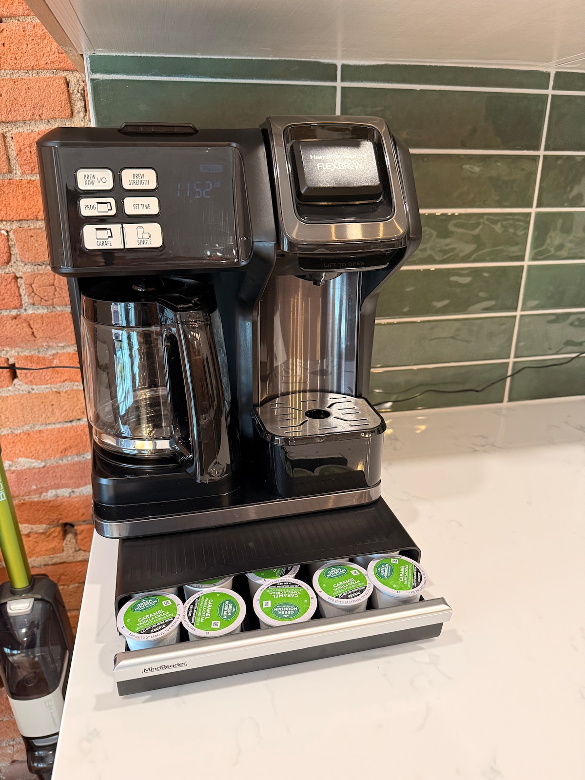 A coffee station featuring a versatile coffee maker with both single-serve and carafe functionality. The machine is black with a digital display and is positioned on a light countertop. Below, a tray holds several single-serve coffee pods, easily accessible for guests.