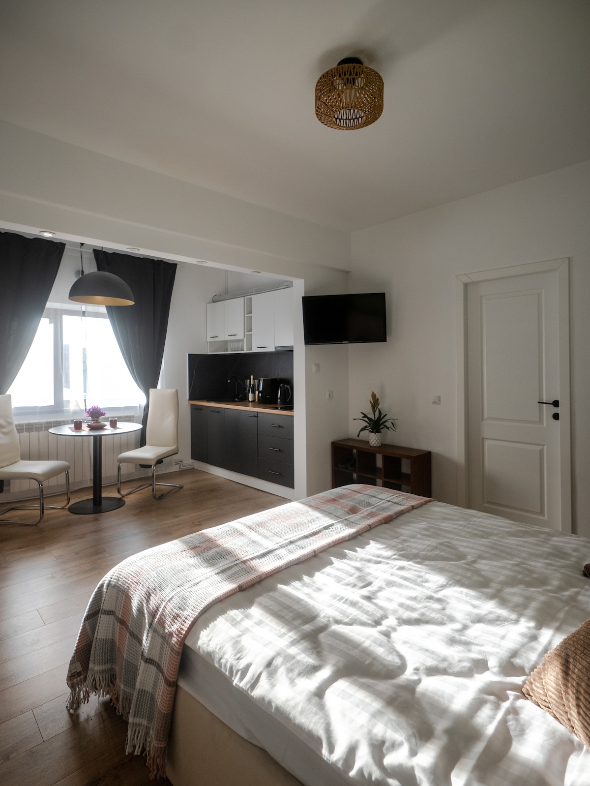 A contemporary room features a spacious bed with a light-colored blanket and soft pillows. A round dining table with four chairs and a small kitchenette is visible. Natural light filters through large windows adorned with dark curtains, enhancing the airy feel of the space.