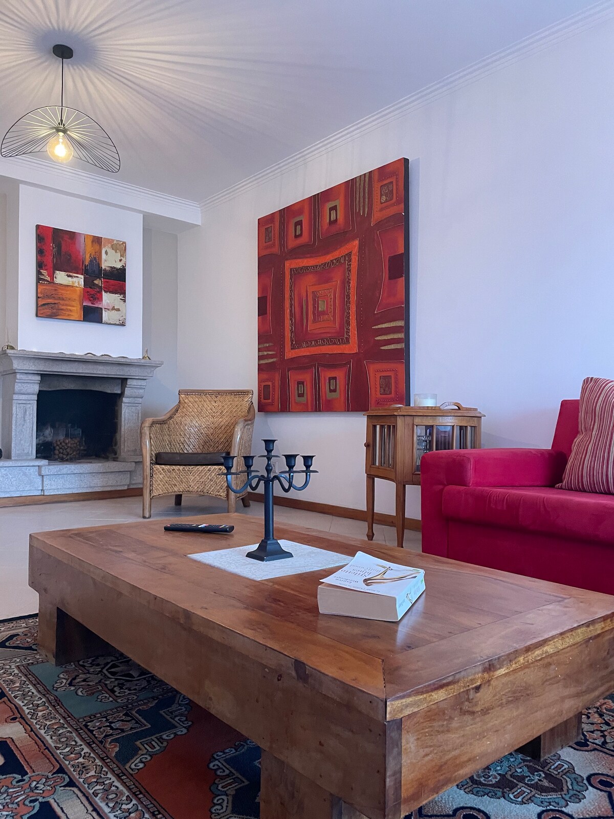 A living area features a wooden coffee table positioned in front of a cozy couch. An artistic wall hanging with vibrant patterns adds a pop of color, while a modern light fixture provides soft illumination. A candle holder rests on the table, complementing the inviting space.