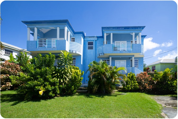 A vibrant blue villa with two separate units is framed by lush greenery and colorful tropical plants. The exterior features white balconies and railings, allowing for an inviting view of the well-maintained garden and clear blue sky.