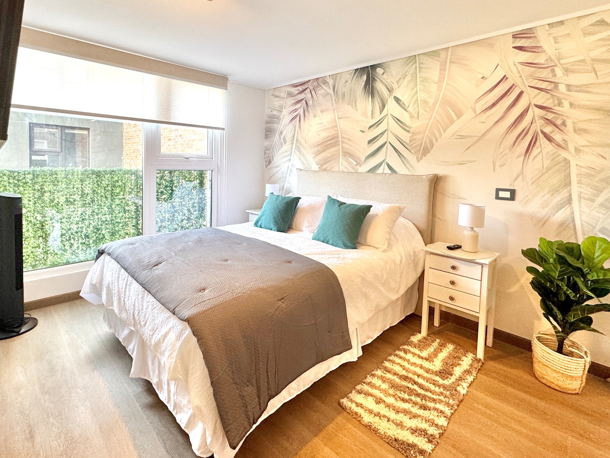A serene bedroom features a spacious bed with light bedding and decorative pillows. A soft rug is positioned on the floor. A bedside table holds a lamp, while large windows provide natural light and views of greenery outside.