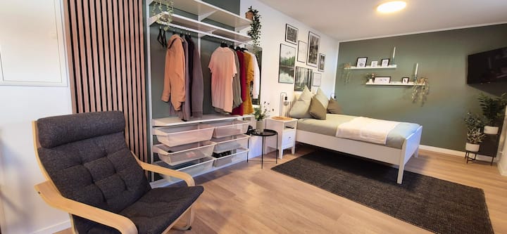 Acogedora sala de estar/dormitorio con armario abierto, acento verde y almacenamiento cuidado, elegante y práctico al mismo tiempo. ✨