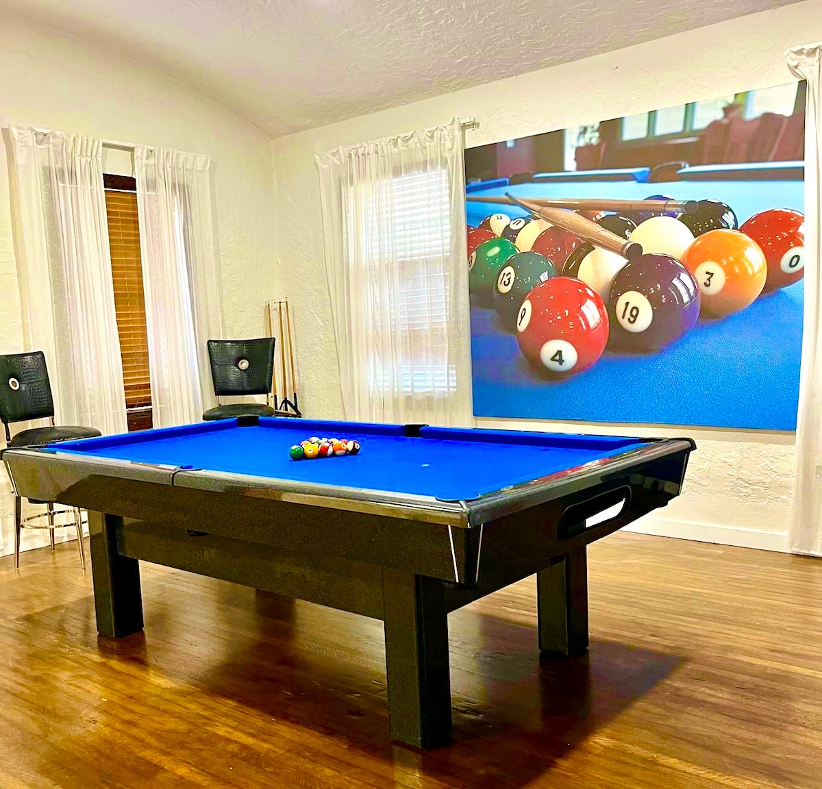 A game room features a vibrant blue pool table, set with colorful billiard balls ready for play. Light wood flooring contrasts with the dark table, while two tall chairs are positioned nearby. Natural light filters through sheer white curtains.