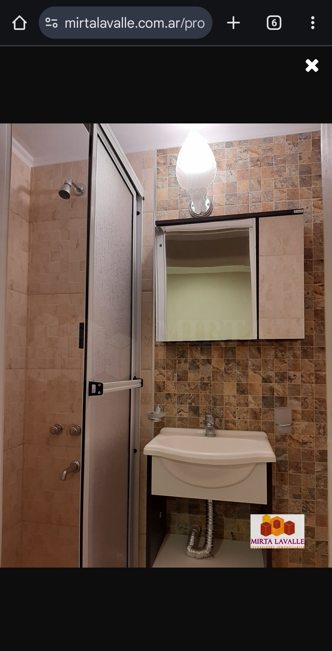 A modern bathroom features a standing shower with a glass enclosure and a sleek sink underneath a mirrored cabinet. The walls are adorned with beige and brown tiles, complemented by a softly glowing light fixture above.