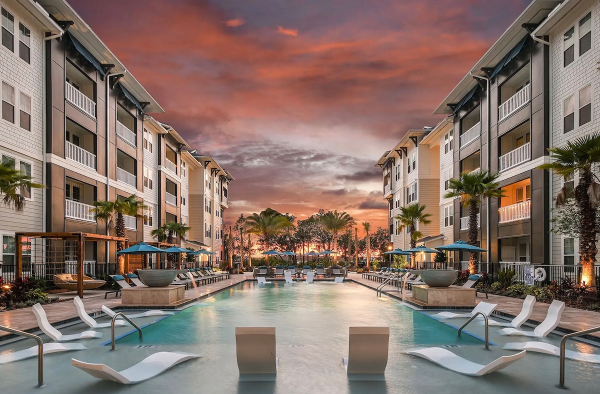 A spacious outdoor pool area is bordered by palm trees and lounge chairs. The modern buildings stand alongside the pool, with their balconies visible above. A vibrant sunset casts warm colors across the sky, enhancing the tranquil atmosphere of the communal space.