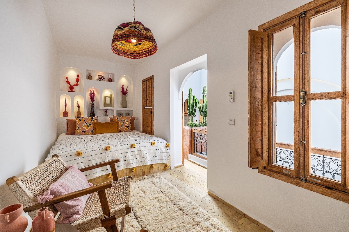 A bright sleeping area features a spacious bed adorned with colorful pillows, complemented by artistic wall niches holding decorative items. A wooden window provides views of the balcony, revealing greenery outside. Textured textiles lay softly on the floor, adding comfort to the serene atmosphere.