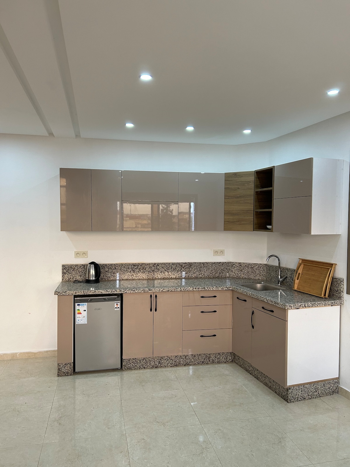 The modern kitchen features sleek cabinetry with a combination of light and dark wood finishes. Countertops in neutral tones provide ample workspace. Stainless steel appliances, including a refrigerator and kettle, are visible. Bright lighting enhances the spacious and functional layout.
