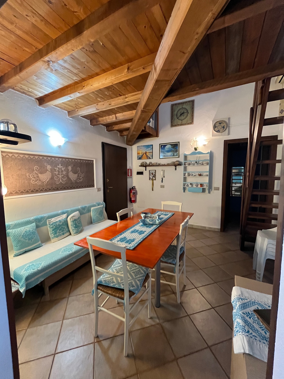 An inviting dining area features a wooden table accompanied by light-colored chairs. A comfortable seating area with patterned cushions is nearby. The ceiling showcases wooden beams, and the walls are adorned with decorative art and a clock, adding warmth to the space.