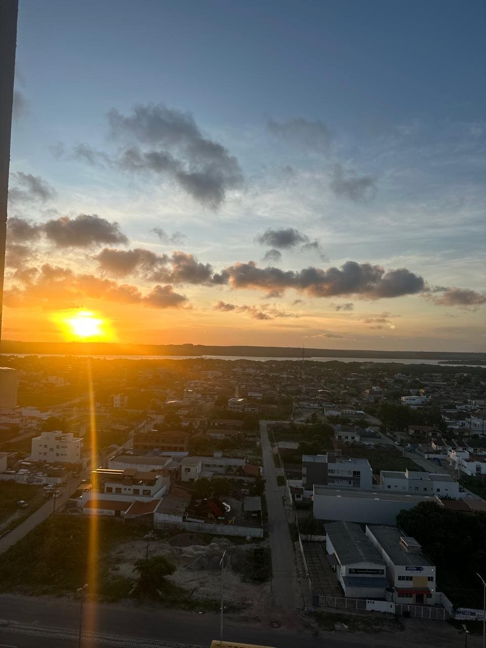 A sunset view is captured from the apartment, showcasing golden rays reflecting off the horizon. The surrounding neighborhood is visible, with buildings lining the streets and greenery peeking through. The sky is filled with soft clouds, creating a serene atmosphere.