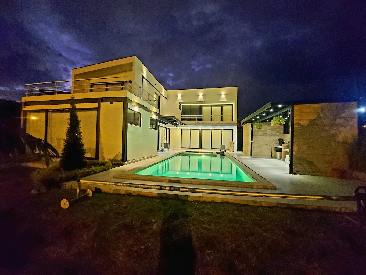 A modern house is illuminated at night, showcasing a clear swimming pool reflecting green hues. Large glass doors and expansive windows allow glimpses of the interior. The well-maintained landscape includes grassy areas and a small tree, enhancing the serene outdoor setting.