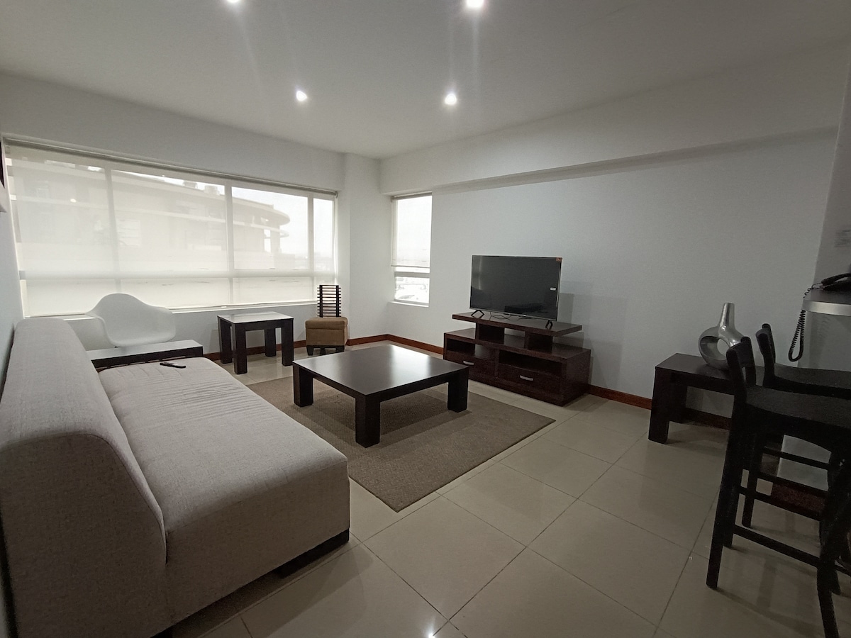 A modern living area features a light-colored sofa and dark wooden furniture, including a coffee table and side tables. Large windows allow natural light to fill the space, while a television is positioned across from the seating area. A dining table with chairs is visible in the corner.