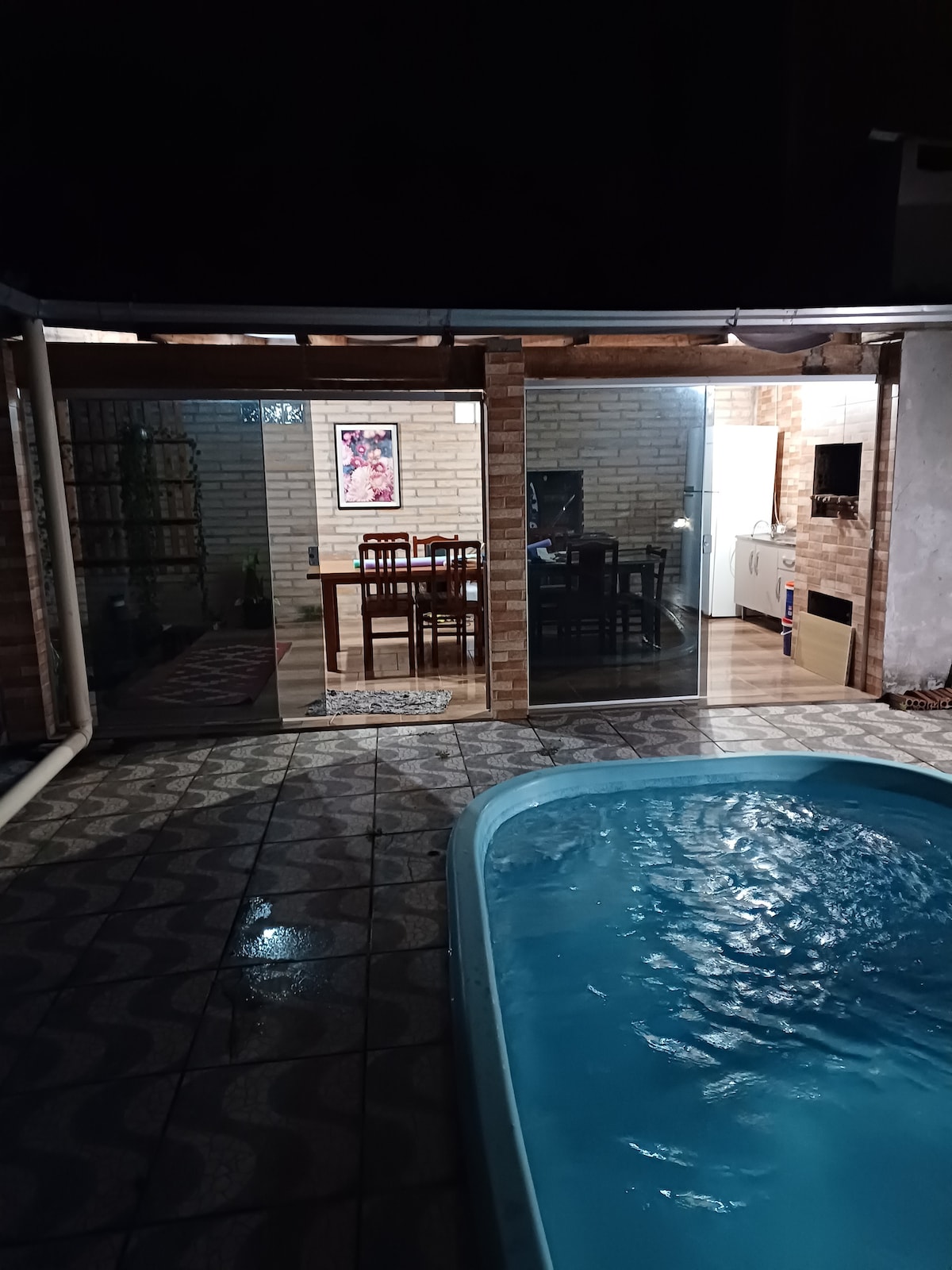 A well-lit area features a pool with a calming blue hue. Glass doors connect the indoor dining space, furnished with wooden chairs and a table, to the outdoor setting. The evening atmosphere is enhanced by the warm glow of lights reflecting off the tiled floor.