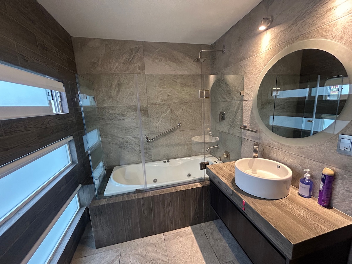 A modern bathroom features a glass-enclosed jacuzzi tub alongside a spacious walk-in shower. Soft lighting highlights the elegant stone tiles and a round mirror above a minimalist sink. Natural light filters through frosted glass windows, adding a touch of brightness to the serene space.