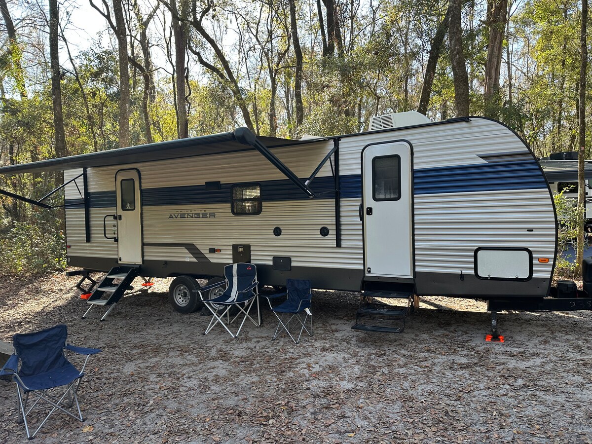 A spacious travel trailer features a streamlined exterior, highlighted by blue and silver stripes. Two foldable chairs are positioned nearby on a sandy surface, and an awning extends from the trailer, providing shade. Surrounding trees create a natural and tranquil setting.
