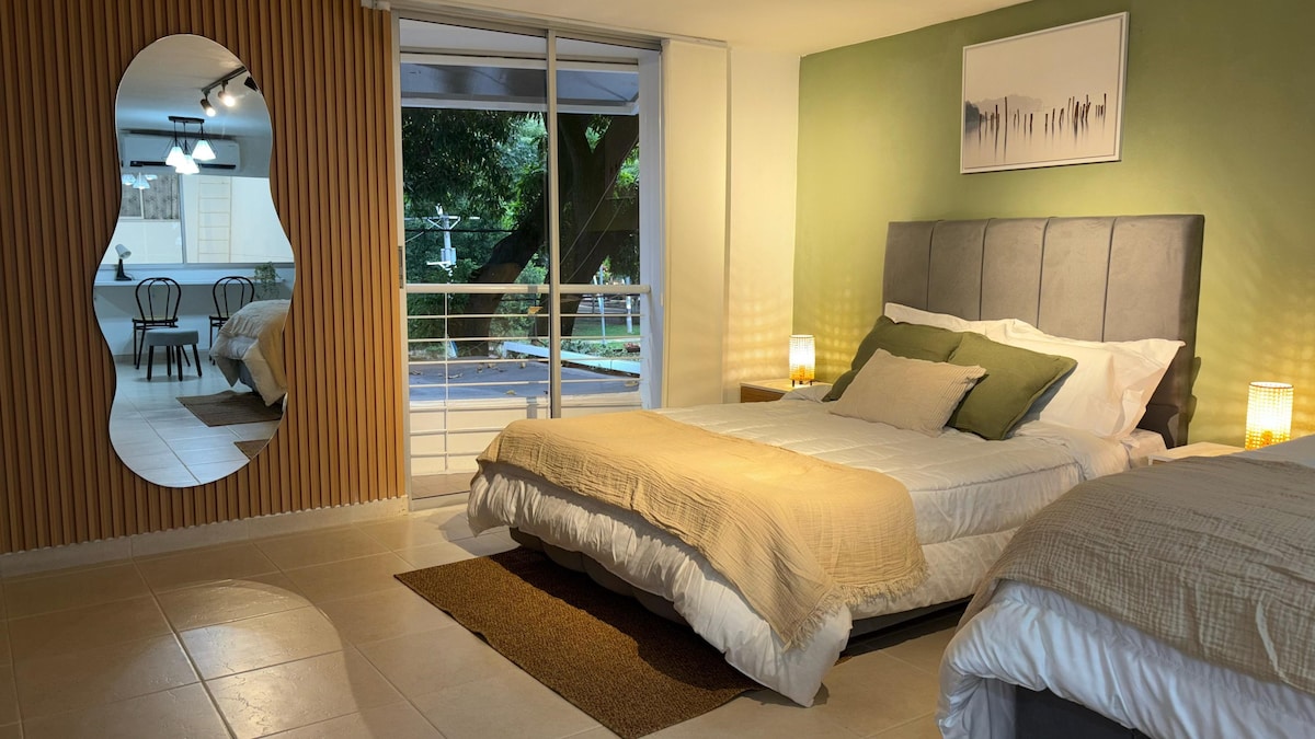 The bright sleeping area features two spacious beds with neutral-toned bedding. Natural light floods in from large glass doors that lead to an outdoor balcony area. A mirror with a unique shape reflects a cozy corner, enhancing the openness of the space.
