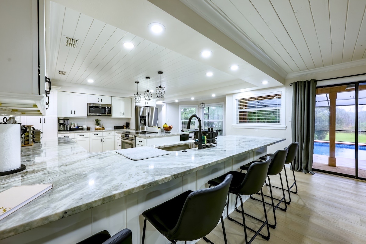 A modern kitchen is showcased with a large island featuring a marble countertop. Several bar stools are positioned at the island, with ample cabinetry and appliances visible. Natural light fills the space through large windows, with a sliding door leading to an outdoor area.