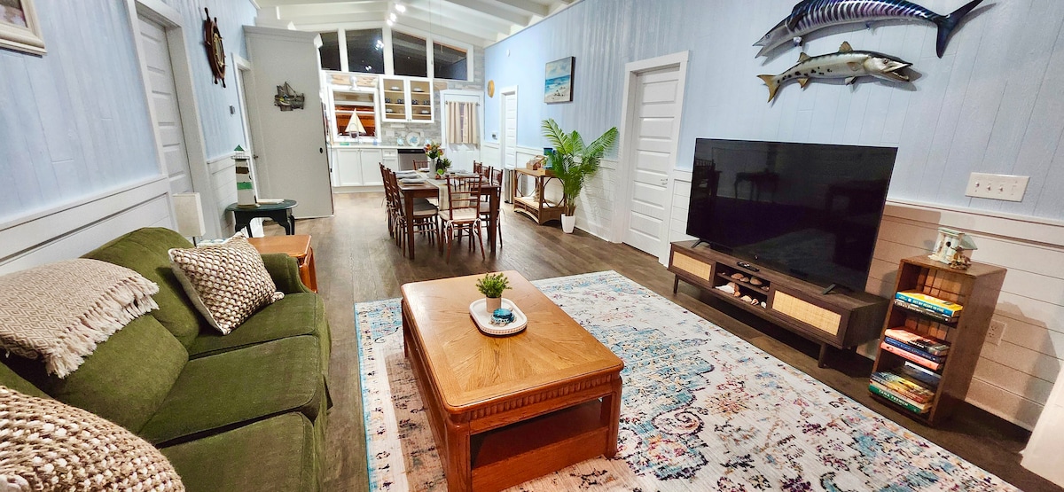 A spacious living area is showcased, featuring a green sofa adorned with textured throw pillows and a large coffee table. A 65" smart TV rests on a media console, while a dining table with multiple chairs is visible in the background, accompanied by bright light from the kitchen area.
