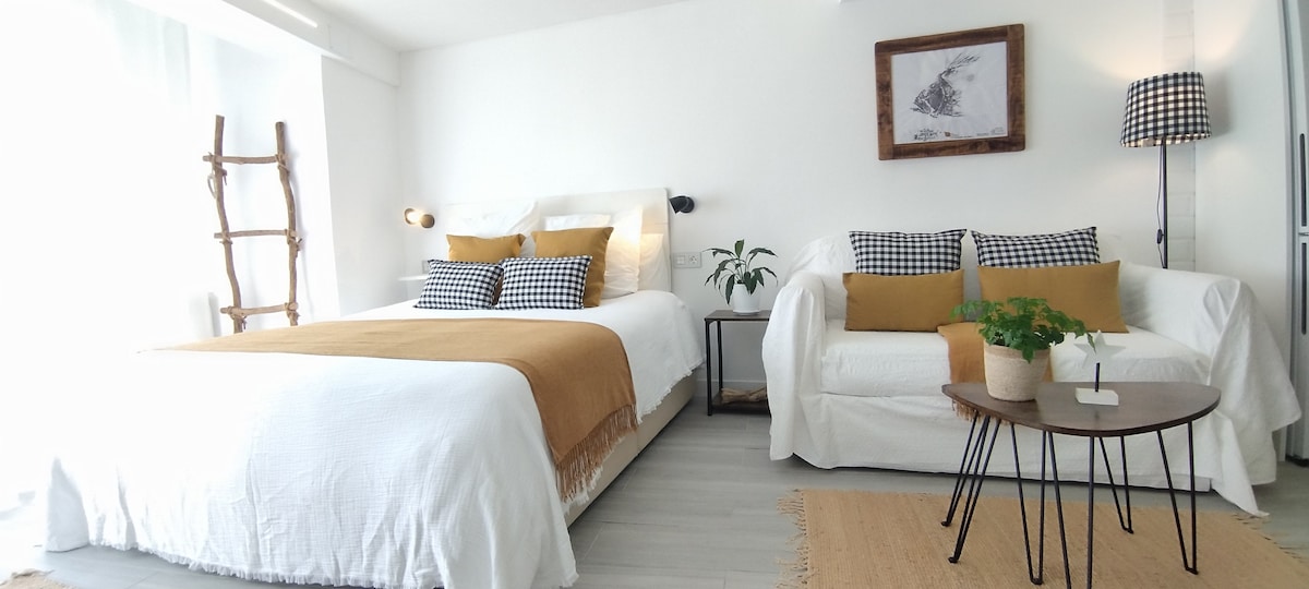 A bright and open space features a spacious bed dressed in white linens, accented by a mustard throw. A cozy sitting area includes a white sofa with patterned cushions and a small wooden table, complemented by natural light flowing through large windows.