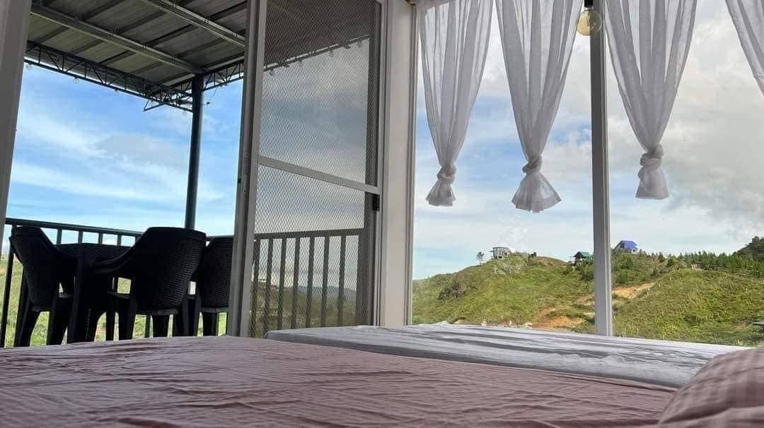 A spacious sleeping area features a soft bed with light bedding. Sheer white curtains frame the open window, allowing natural light to illuminate the room. A patio with chairs is visible beyond, offering views of the surrounding landscape.