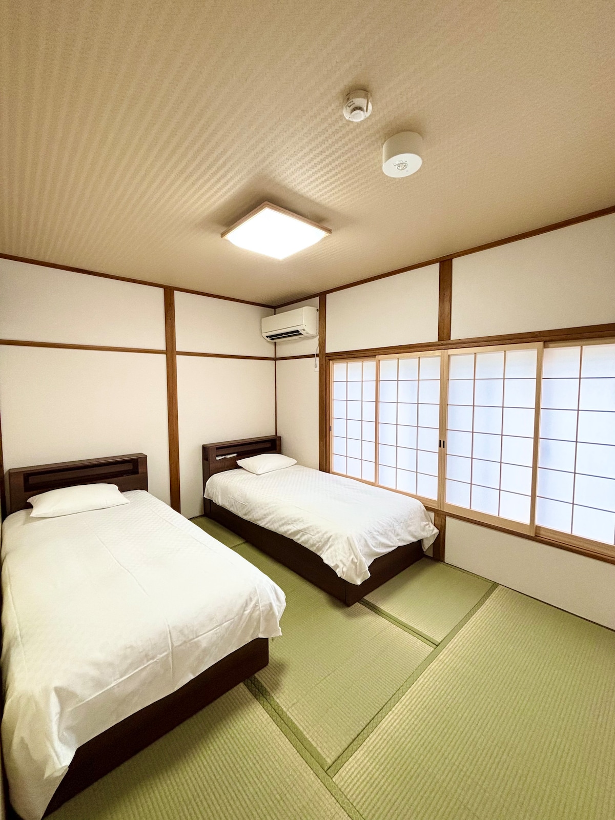 A traditional tatami bedroom features two twin beds with crisp white linens. Natural light enters through a wooden-framed shoji screen, illuminating the warm tones of the room. Wall panels are light-colored, and a ceiling-mounted light provides additional brightness.