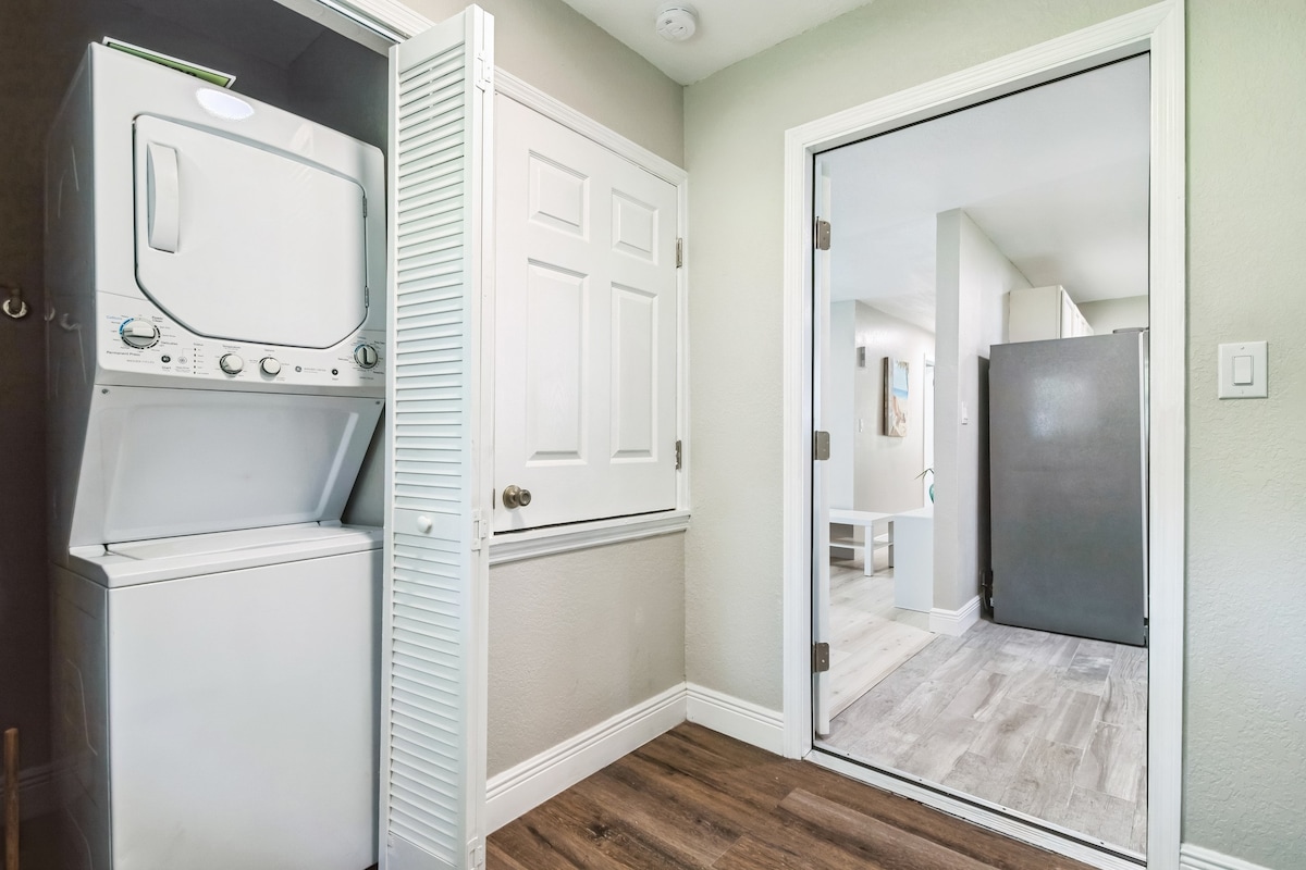The laundry area features a stacked washer and dryer unit, conveniently located near a door leading to the living space. The grey walls and natural wood flooring create a clean atmosphere, and the nearby door provides a view into the home's interior.
