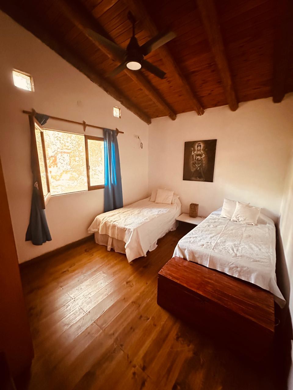 A simple bedroom is furnished with two twin beds, topped with white linens. A wooden trunk is placed at the foot of one bed. Natural light enters through a window adorned with blue curtains, and the room features a wooden ceiling and floor.