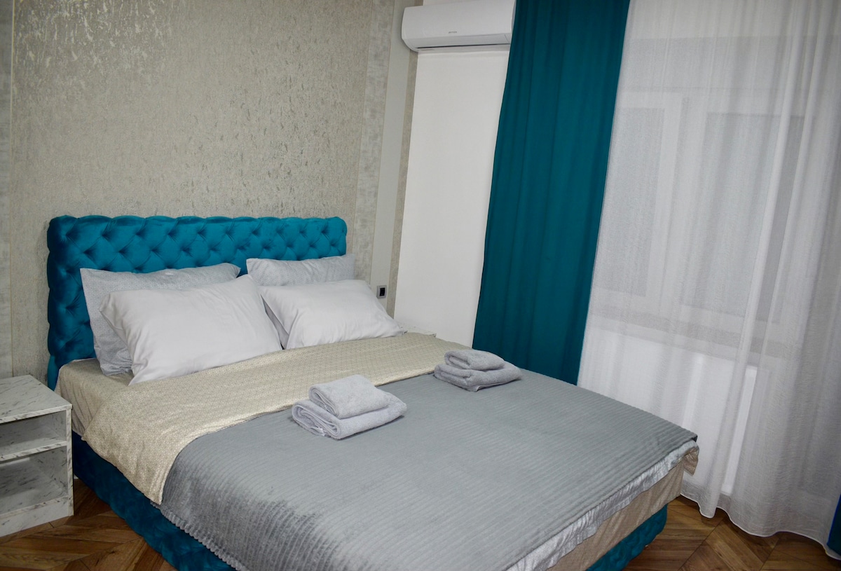 A spacious bed with a tufted blue headboard is presented, complemented by white pillows and a light grey blanket. Two rolled towels are placed neatly at the foot of the bed. Sheer curtains allow natural light to brighten the room, while a textured accent wall adds visual interest.