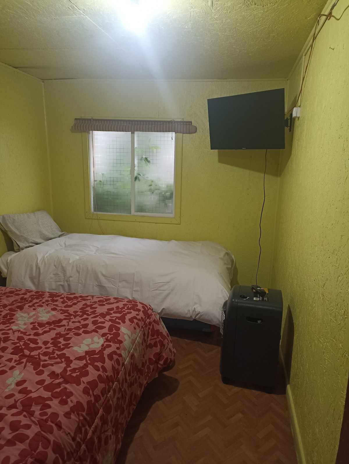A cozy room features two beds, one with a floral-patterned comforter and the other with a white cover. A small television is mounted on the wall opposite the beds. A window with light sheer curtains allows natural light in, showcasing greenery outside.