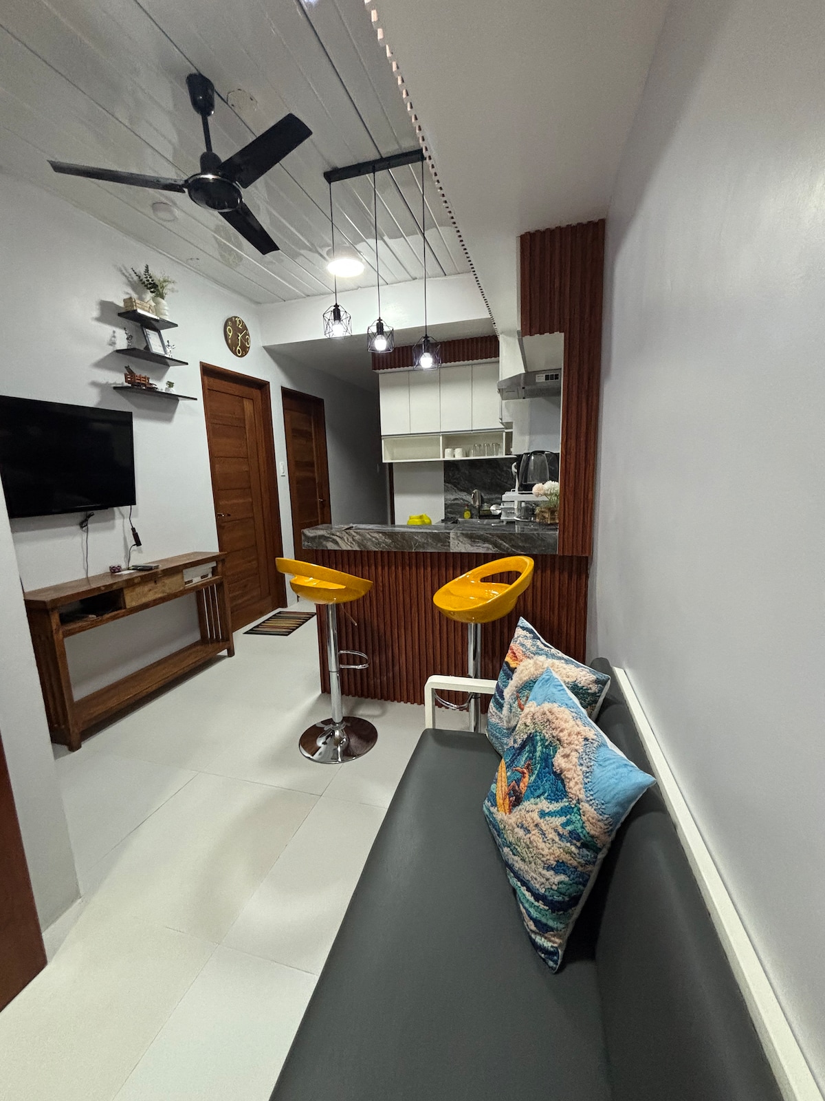 A modern living area features a comfortable sofa adorned with a decorative pillow. Two high stools in bright yellow are positioned at a small kitchen counter. A television is mounted on the wall opposite, and a ceiling fan offers a cooling presence.