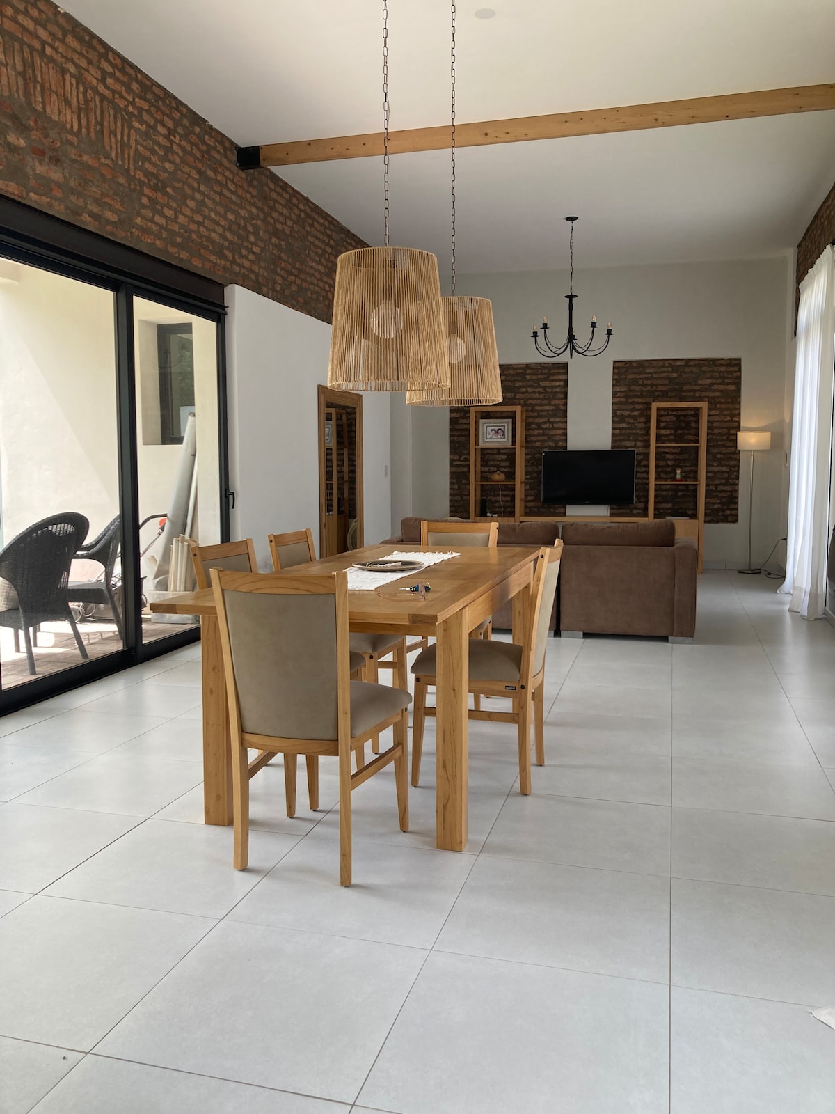 A spacious dining area features a wooden table surrounded by six chairs, with light filtering through large glass doors. Two woven pendant lamps hang above the table, and a comfortable seating area with a sofa is visible in the background.